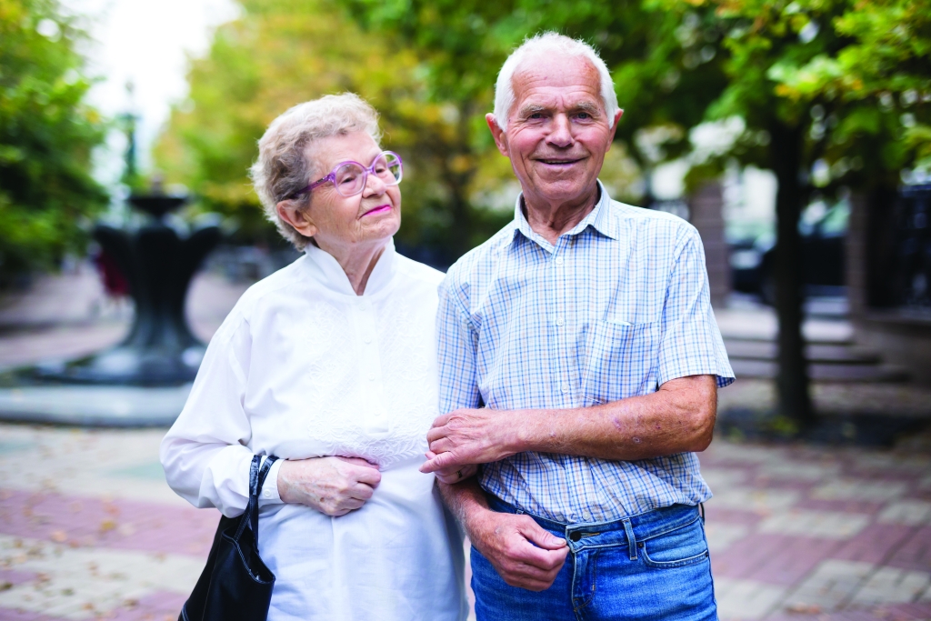 a man and a woman walking in the park with their arms linked. They appear to be in conversation and they are smiling.