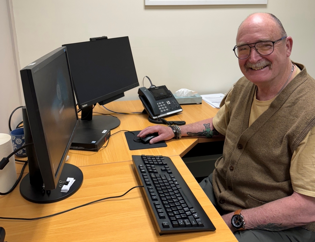 Trevor is an older gentleman pictured sitting at his desk. He is smiling.