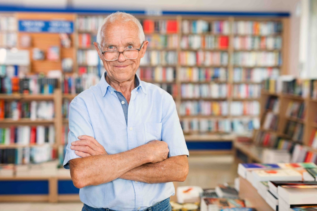 An older man smiles at the camera with his arms folded. He looks like he is in a public library.